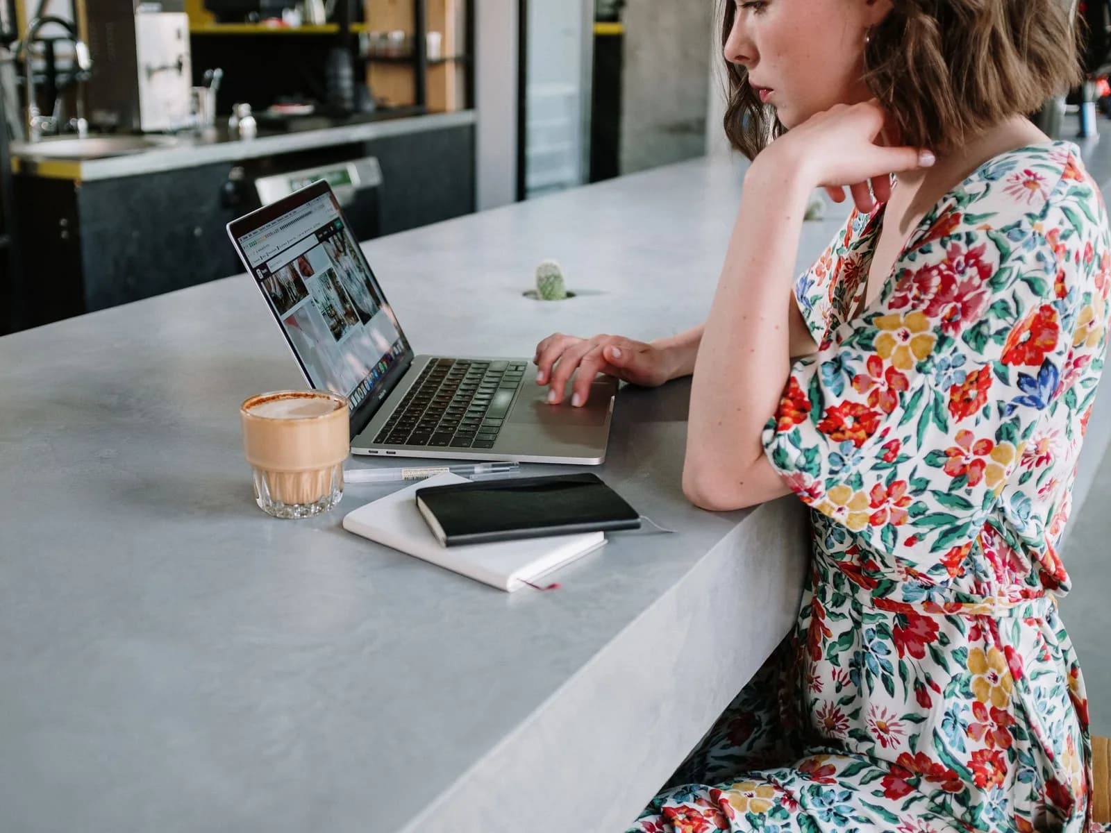 Mulher de vestido às flores ao portátil, com um café e dois cadernos no balcão